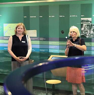 Zwei Frauen stehen in einer Ausstellung; eine spricht ins Mikrofon